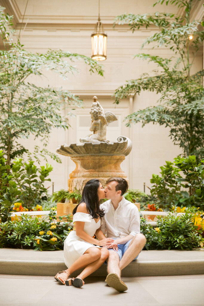 A man and a woman sit at the edge of a fountain, kissing, surrounded by plants