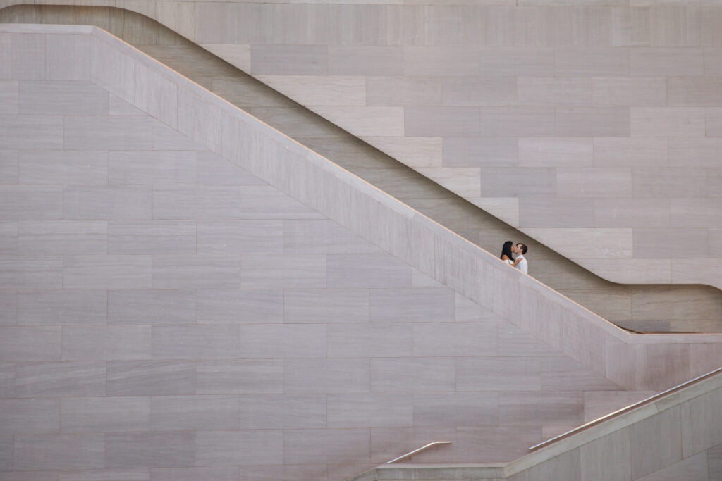 A photo taken from afar of a man and a woman kissing on the National Gallery of Art staircase, surrounded by white marble