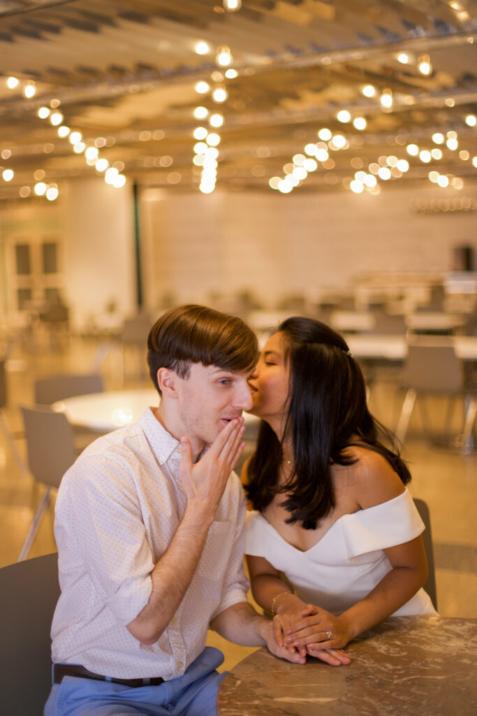 A woman whispers in a man's ear, who makes a joking shocked face. They are illuminated by string lights in the back