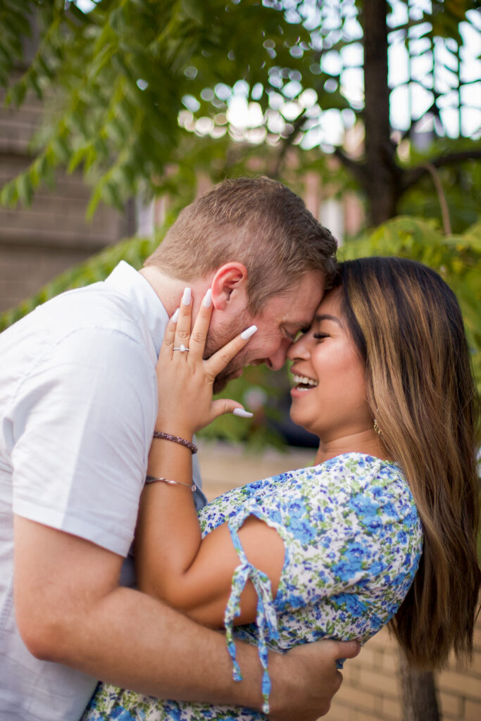 A man and woman laugh together as the woman's hand on the man's cheek displays her engagement ring